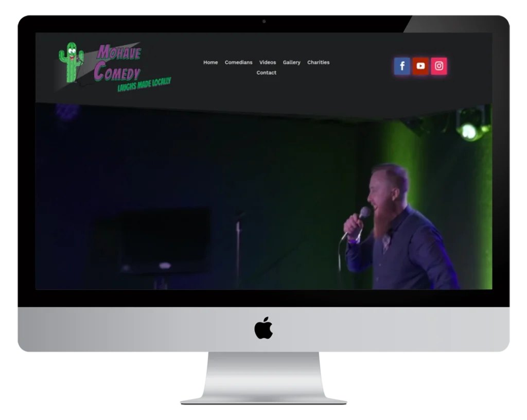A desktop computer displays a website for Mohave Comedy, featuring a header with navigation links and a background image of a bearded man performing stand-up comedy with a microphone.