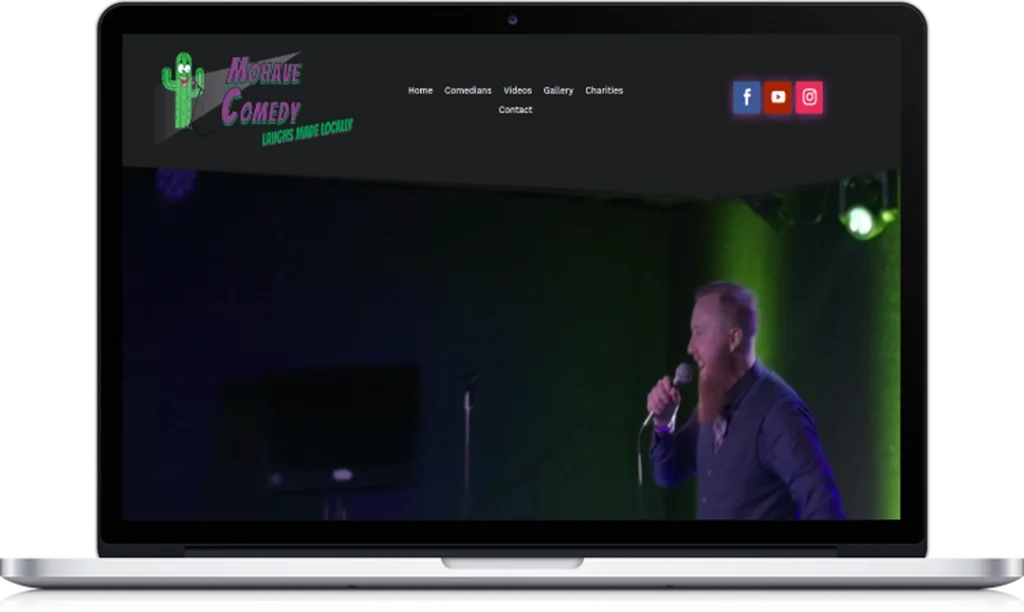 A laptop displays a website featuring a stand-up comedian performing on stage with a microphone. The website header reads 
